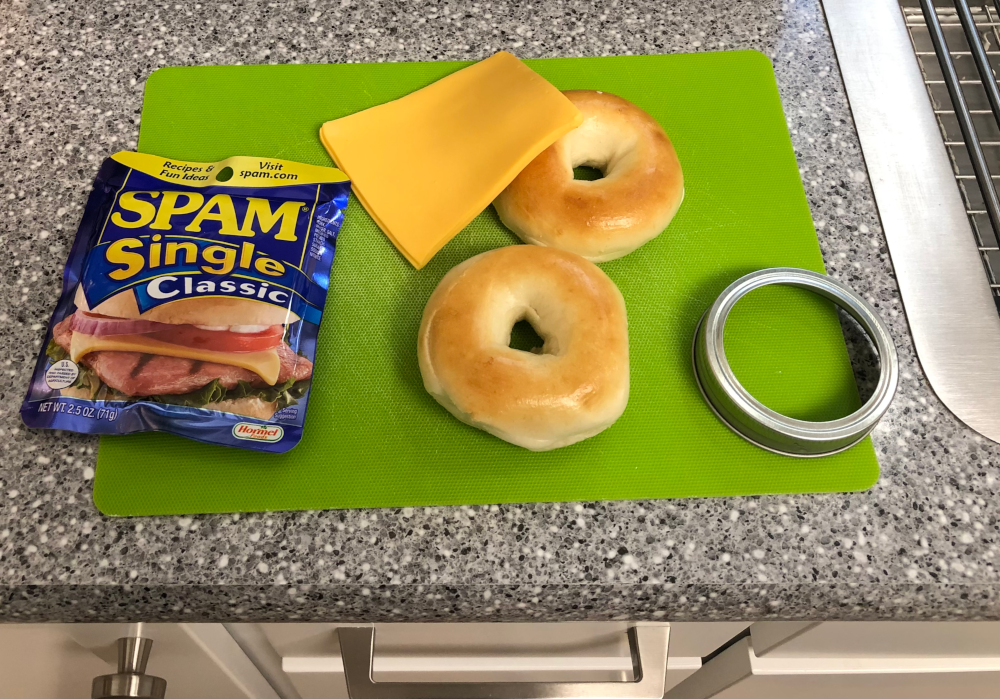SPAM Single package on a green cutting mat. Also on the mat are two slices of yellow/orange American cheese, two plain mini bagels, and the screw-ring for a half-pint canning jar.