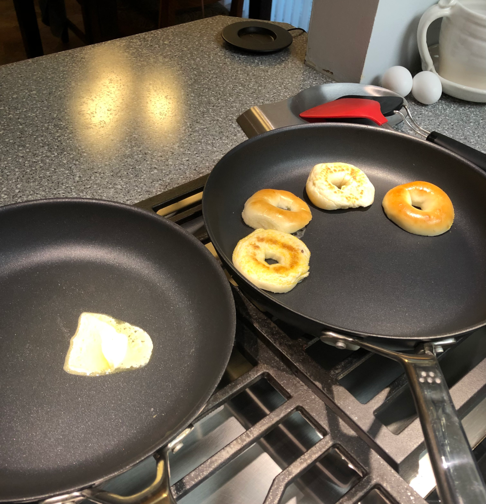 Two frying pans on a gas cooktop. One has butter melting, the other has 4 halves of plain mini bagels on it, cut-side down, to toast just the inside surface. In the background, a couple spatulas and a couple eggs.