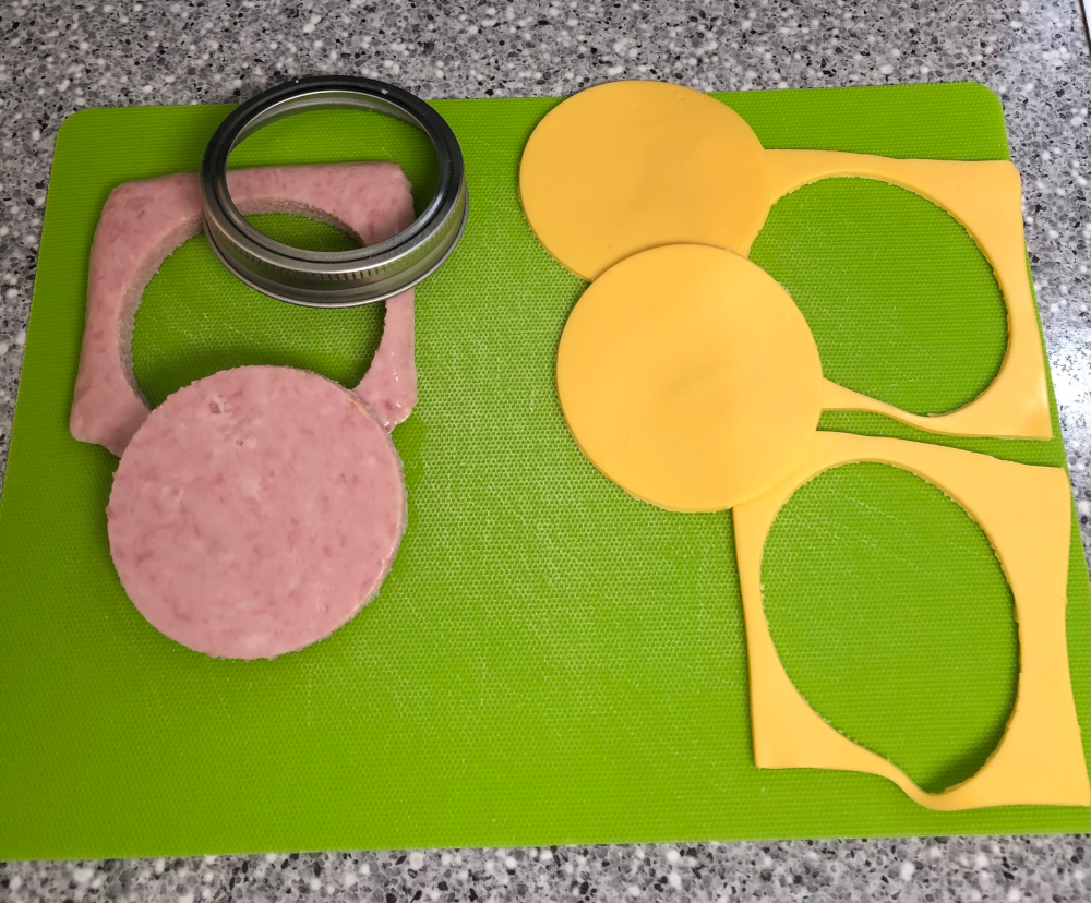 The green cutting mat again. But now the SPAM has been cut into a circle by using the canning ring like a cookie cutter. The slices of cheese have been separated from each other, and also each cut into circles. The off-cuts with holes in them are also sitting on the cutting mat.