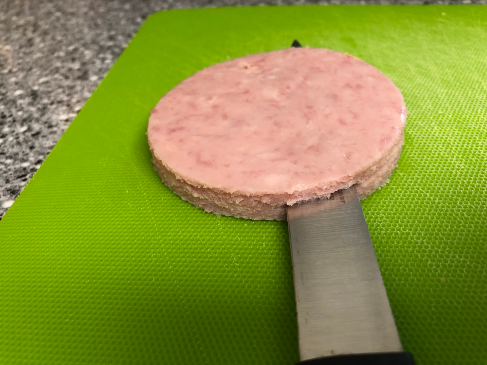 Close-up of the circle of SPAM, being sliced to half-thickness with a fine knife blade.