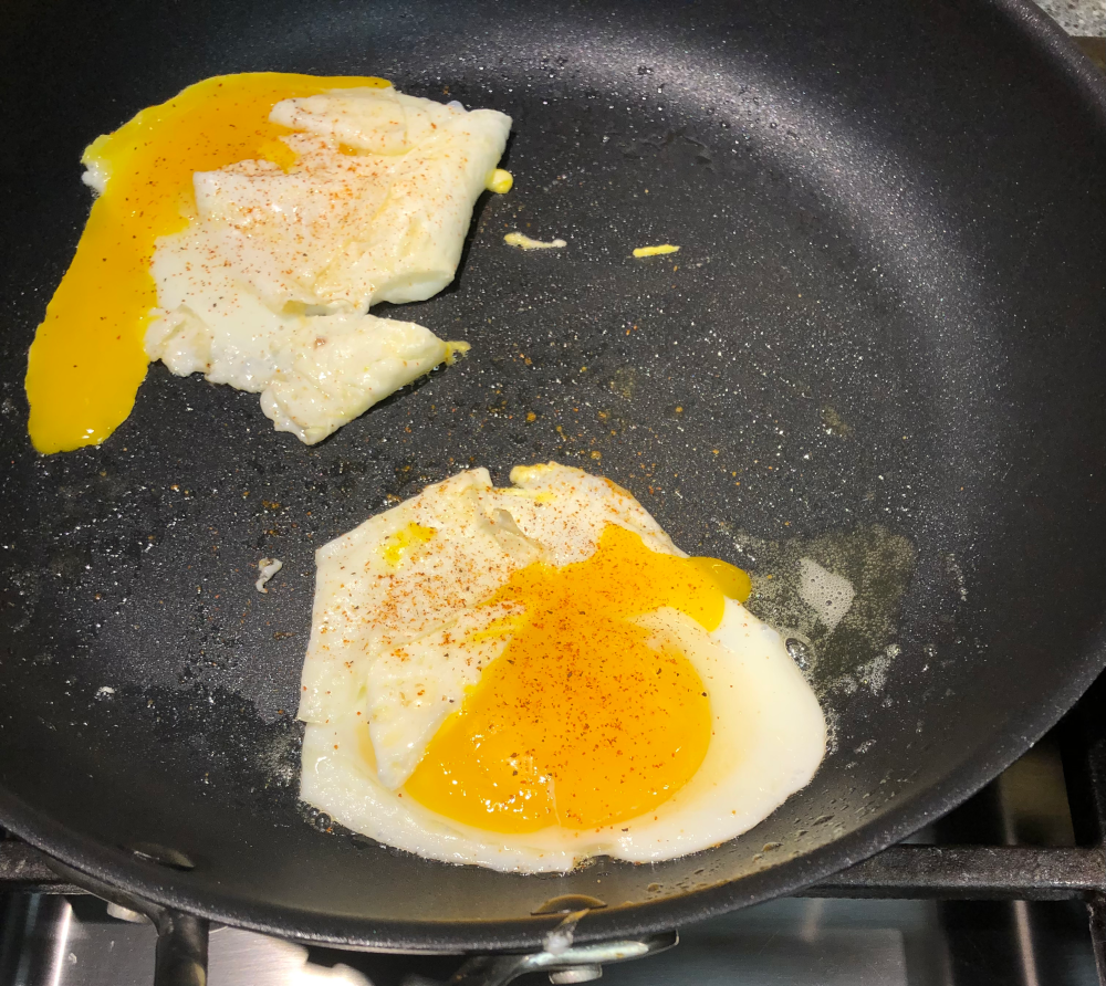 Same two eggs frying in a pan, but now they have spices sprinkled on them.