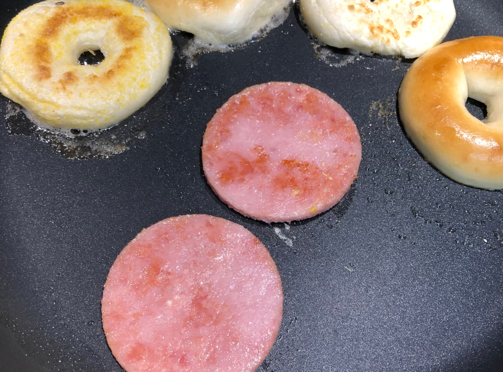 Closer photo of the SPAM circles, now having been frying in the same pan as the bagels, and have been flipped once. The surface of the SPAM has patches of brown.