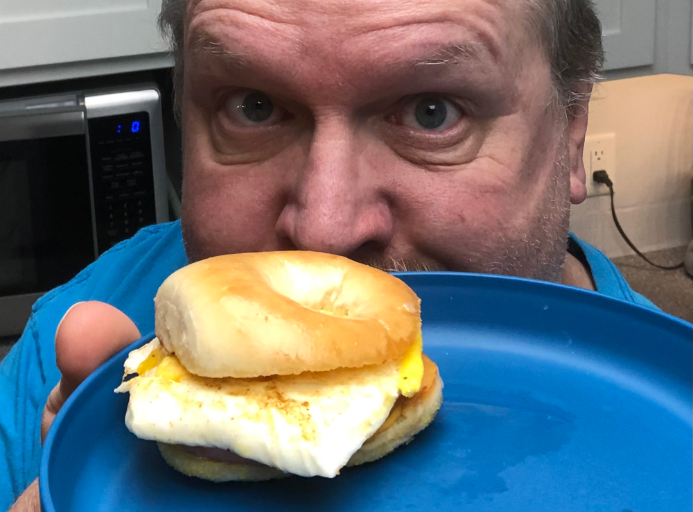 A big fat weirdo peers giddily from behind a blue plate with a breakfast slider on it. He looks very pleased, but somewhat unhinged. The slider looks delicious, though, if you're into that sort of thing.
