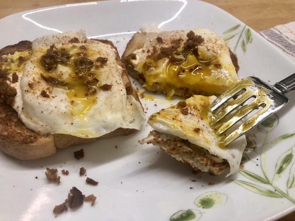 White plate with white toast, over-easy eggs, and bacon bits. A fork holds a cut-off bite, and the tangs of the fork show liquid yolk detritus from the cutting and poking of the next bite. That is, it is immediately apparent that the eggs were not over-done.